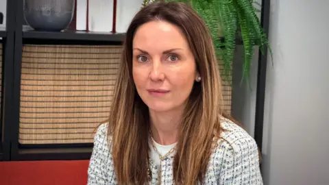 Solicitor Laura Connor in her office. She has long, brown hair and is wearing a white and blue cotton jacket. There is a shelf behind her with a plant on it