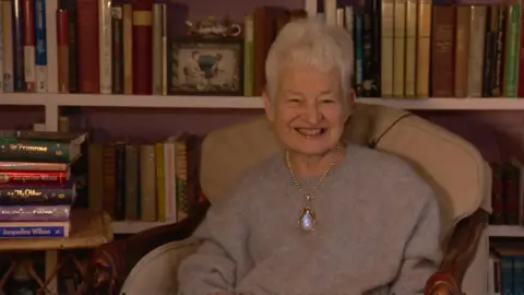 Jacqueline Wilson sits in an armchair in front of bookshelves