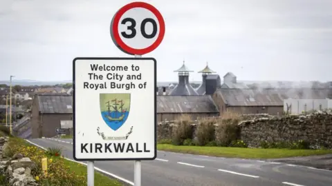 Sign which says 'Welcome to The City and Royal Burgh of Kirkwall' with an image of a tall ship, with roofing of the town in the background.