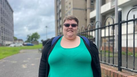 Woman stands outside high rise flats wearing green top