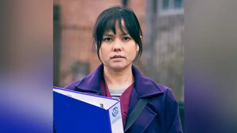 ITV/Coronation Street Woman cast-member wears a purple jacket and carries a purple folder.