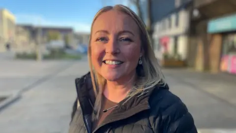 A woman with mid-length blonde hair, wearing a black puffer coat on the shopping street. It was a head and shoulder shot. He is in focus and the background is blurred.
