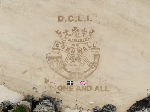 Rocks Up The badge of the Duke of Cornwall's Light Infantry drawn in the pale sand. The badge features a crown, the word 'Cornwall', two thistles and a horn. The Cornish flag and UK flag lie on the sand below it. 