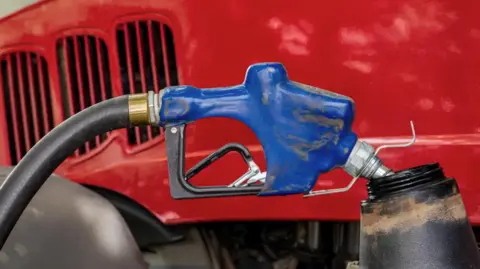 Getty Images Photograph of a blue diesel pump resting on the fuel cap of a red tractor. The pump and the fuel cap are slightly splashed with mud.