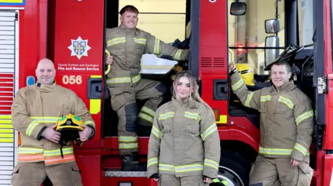 Three men and a woman stood in fire fighter uniform in front of a fire engine. One of the men is stood on the step of the fire engine.