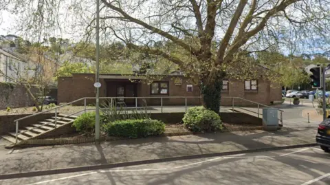 The front of Newton Abbot Magistrates' Court. It is a modern single-storey brick building with a road and a large tree in front of it.