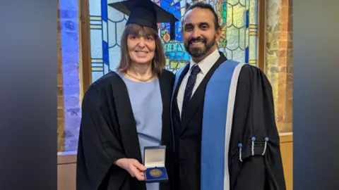 A woman is wearing a black university cloak with a pale blue dress underneath. She is holding a medal in a presentation case in her right hand. She has brown shoulder-length pale brown hair and brown eyes. She is wearing a mortar board on her head. To her left is a man who has a dark beard and short dark hair. He is wearing a university cloak which is black and has a band of pale blue and white to the edge. He has a dark suit, white shirt and blue tie underneath.