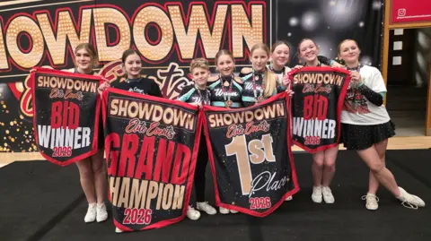 South Coast Waves cheerleaders Eight young people gathered around several first placed sign. They are wearing leotards and in front of some huge branding. They are at a competition with lots of people milling around behind.