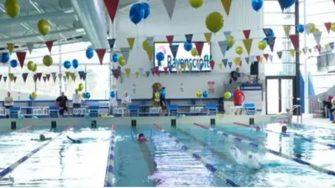 BBC A swimming pool with swimmers doing laps under bunting and balloons.