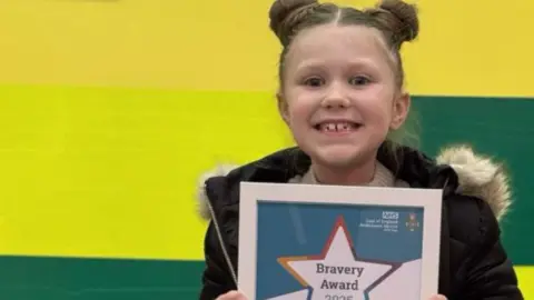 A little girl grinning while holding a framed certificate in front of her mum and the call handler, behind all of them is a bright yellow and green ambulance