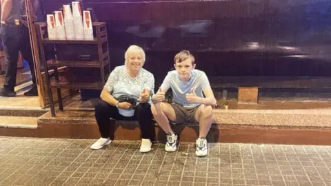 Gail Iles Gail Iles and Mason sitting on a step together. Mason is giving a double thumbs up.