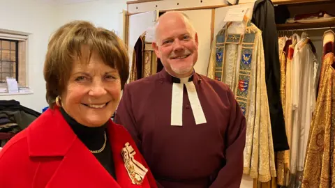 Jon Wright/BBC Clare Euston, in a red coat, and The Very Reverend Joe Hawes in clerical attire, stand smiling at the camera.