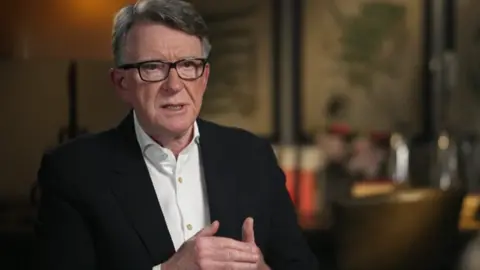 Lord Mandelson sits in a chair while being interviewed. He is wearing a white shirt and a black blazer. He is also wearing glasses and has greying dark hair, styled in a side parting. Also visible is the blurry background of what looks to be a modern looking room.