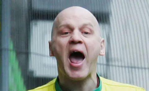 Michael Stone shouts open-mouthed at the camera. He is wearing a yellow top.
