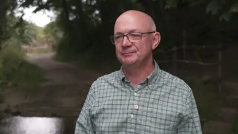 A bald man, wearing glasses and a check green shirt, standing in front of a river. 