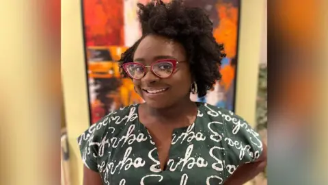 Seyi B Seyi is wearing a dark green blouse and smiling at the camera. She has big red glasses frames and neatly styled hair.