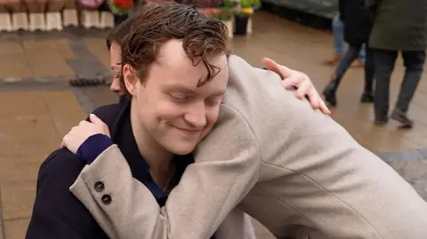 Martin Giles/BBC Rodney-Jones is sitting down outside, wearing a navy coat and has brown and slightly ginger hair. Laura Leate has brown hair, wearing a light brown coat, and she is leaning over him as she hugs him. In the background are paving slabs and some potted flowers arranged in front of a market stall.