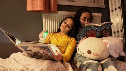 Family Amina and Hajar are tucked up with their books at bedtime under a duvet next to a radiator with cuddly toys. Amina is shining a torch on the page. The sisters are cuddled up together.