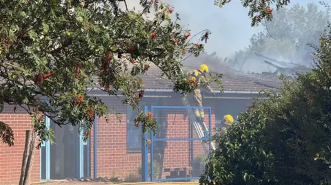 Jamie Niblock/BBC A fire officer is half-way up a ladder leant against a one-storey building wall and roof. Another officer is on the ground looking towards the person on the ladder. Some double doors are open and there is shrubbery and a tree in the front of the building.