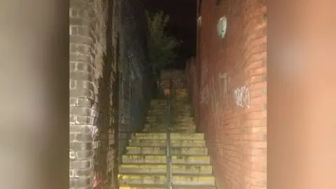 Dark alleyway with a slight flash from the camera. There's a handle and yellow safety taps on the end of each step. The lights are on the right hand bricked wall and are off.