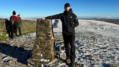 HOWARD KNIVETON A man stands on a hill top, there is a light dusting of snow. He leans on a landmark made from a pile of stone. Two other people look away at the expansive view against clear blue sky.