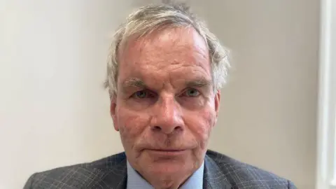 Sharon Edwards/BBC A close-up portrait of a man looking directly into the camera with a serious expression. He has short grey hair and is wearing a grey blazer and light blue shirt. The wall in the background is white.
