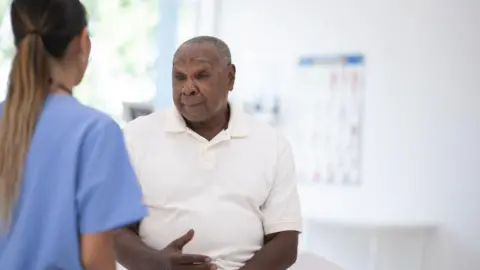 An older black men wearing a white shirt sits on a bed in a clinic talking to a nurse dressed in a blue uniform.