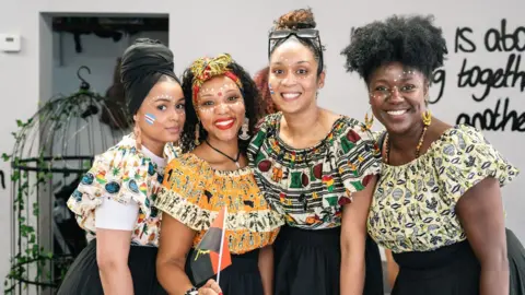 The image shows four people posing side by side for a group photograph inside a modern indoor space. Each person is dressed in colorful patterned outfits featuring geometric and illustrative prints in shades of orange, green, cream, black, and yellow. Several people have white decorative face paint applied in dotted patterns across the forehead, cheeks, and nose area. 