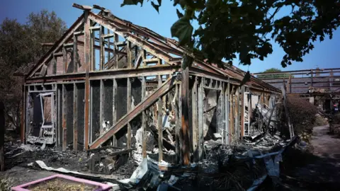 The charred remains of a collapsed hospital building stand amid scattered debris, with blackened wooden beams exposed.