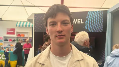 Osian Morgan has brown hair. He is wearing a cream shirt over a white T-shirt