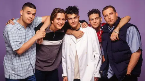 Getty Images All five band members pose in front of a purple backdrop. They each have their arms around each others shoulders casually and smile towards the camera. 