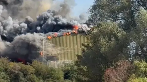 Lee Curtis A green metal building is on fire. Large flames and thick grey smoke are coming from the building. There are several trees and bushes in the foreground.
