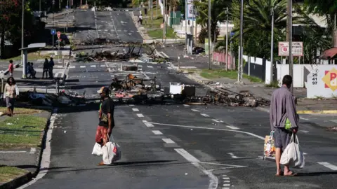 Getty Images People walk past a roadblock barricade set up by rioters near a supermarket, which has reopened to allow people to buy groceries and food, in Noumea, in the French Pacific region of New Caledonia, on May 18, 2024