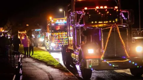 The Wolds Truck and Tractor Run A close up of the cab of a large truck adorned with Christmas lights. Four trucks and tractors, also lit up, follow behind it. Spectators can be seen on a pavement at the side of the road.