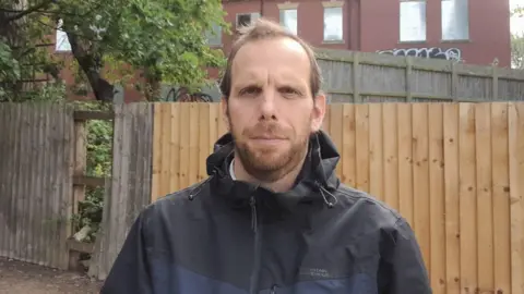 A man with short brown hair and a beard is wearing a dark-coloured raincoat. Behind him a wooden fence, a section of which is missing. There is a tree behind part of the fence and a building which appears to be boarded up.