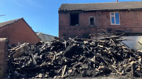 BBC/Victoria Scheer The brick house behind debris shows clear signs of fire damage. One upper-floor window opening is completely empty, revealing a dark, soot-covered interior, while a smaller adjacent window also appears damaged with blackened edges. The brickwork beneath and around the windows is heavily stained with smoke and scorch marks, creating dark patches against the red brick. The roof tiles remain in place, but the eaves above the damaged window look discoloured and uneven.
