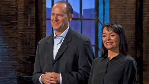 BBC Studios Antony and Lisa Hicks stand side by side in the Dragon's Den to pitch to the show's entrepreneurs. Antony has a black suit with a blue shirt underneath and Lisa is next to him smiling with a black shirt on.