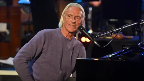 Paul Weller is sat at a piano, smiling. He has chin length blond hair and is wearing a purple jumper. 