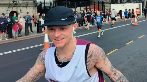 Jack Taylor running the London Landmarks Half Marathon last year. He is wearing a black and white top and has a baseball cap and headphones. There are crowds on the side of the road.