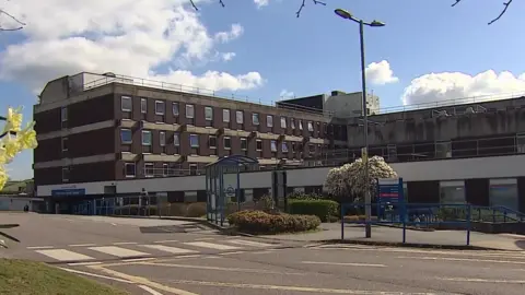 A view of outside North Devon District Hospital. The building is brown with four floors and each floor has many windows. There is a zebra crossing to the left, leading to the hospital.