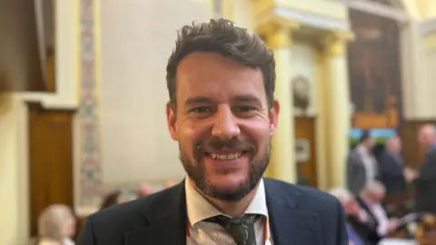 Elliot Deady/BBC A man with a beard smiling straight at the camera. He is wearing a dark suit with a white shirt and patterned tie. Several people are standing and sitting in the background.