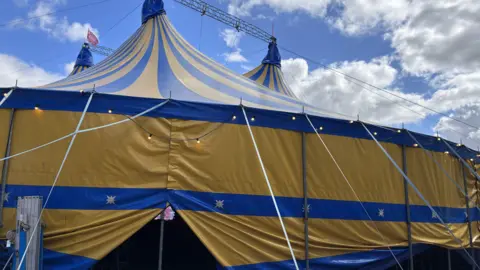 A yellow and blue tent with three pointy peaks. The sky is blue with some light cloud and the photo has been taken from ground level looking up.