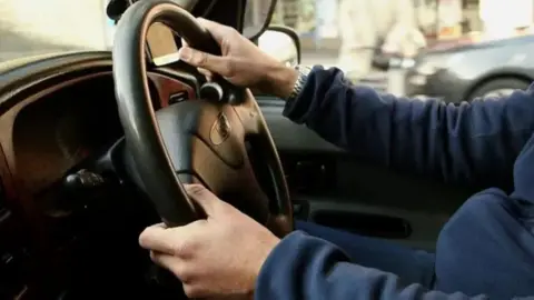 The hands of a person gripping a black steering wheel. They are wearing a silver watch on their right wrist.