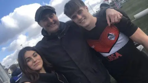 Family photo A woman with dark hair and wearing a black coat, a man wearing a black coat and cap, and a man wearing a black rugby shirt, smiling at the camera with their arms round each other.