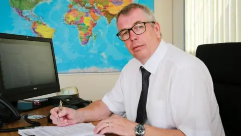 Simon Boyd Simon Boyd wearing a white shirt and dark tie, sat at a desk and holding a pen as he write on paper, with a computer and a map behind him
