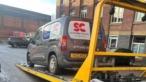 A grey van advertising sports coaching services being hauled onto a ramp and taken away outside Meadow Mill apartments in Stockport. 