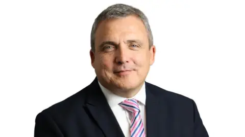 Mark Axcell, incoming chief executive of Nottinghamshire Healthcare NHS Foundation Trust. He is a white man with greying short hair. He is wearing a white collared shirt, black blazer and purple, white and blue striped tie.
