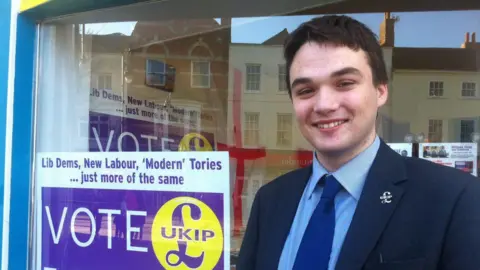 A younger Robin Hunter-Clarke standing outside an office. In the window is a UKIP poster. He is wearing a dark blue suit, lighter blue shirt and a blue tie - with a UKIP badge on his left lapel.