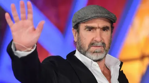 Getty Images Eric Cantona in a flat cap and suit, waving 