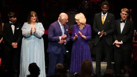PA Media The monarch is dressed in what appears to be a navy blue suit, stood next to his wife, who is wearing a pattered purple dress. They are flanked by award winners who are in dinner jackets